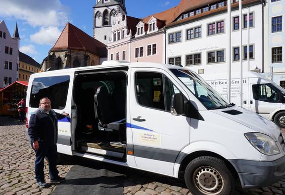 Mann um die 60 an weißen Minibus gelehnt lächelnd vor Stadtkulisse